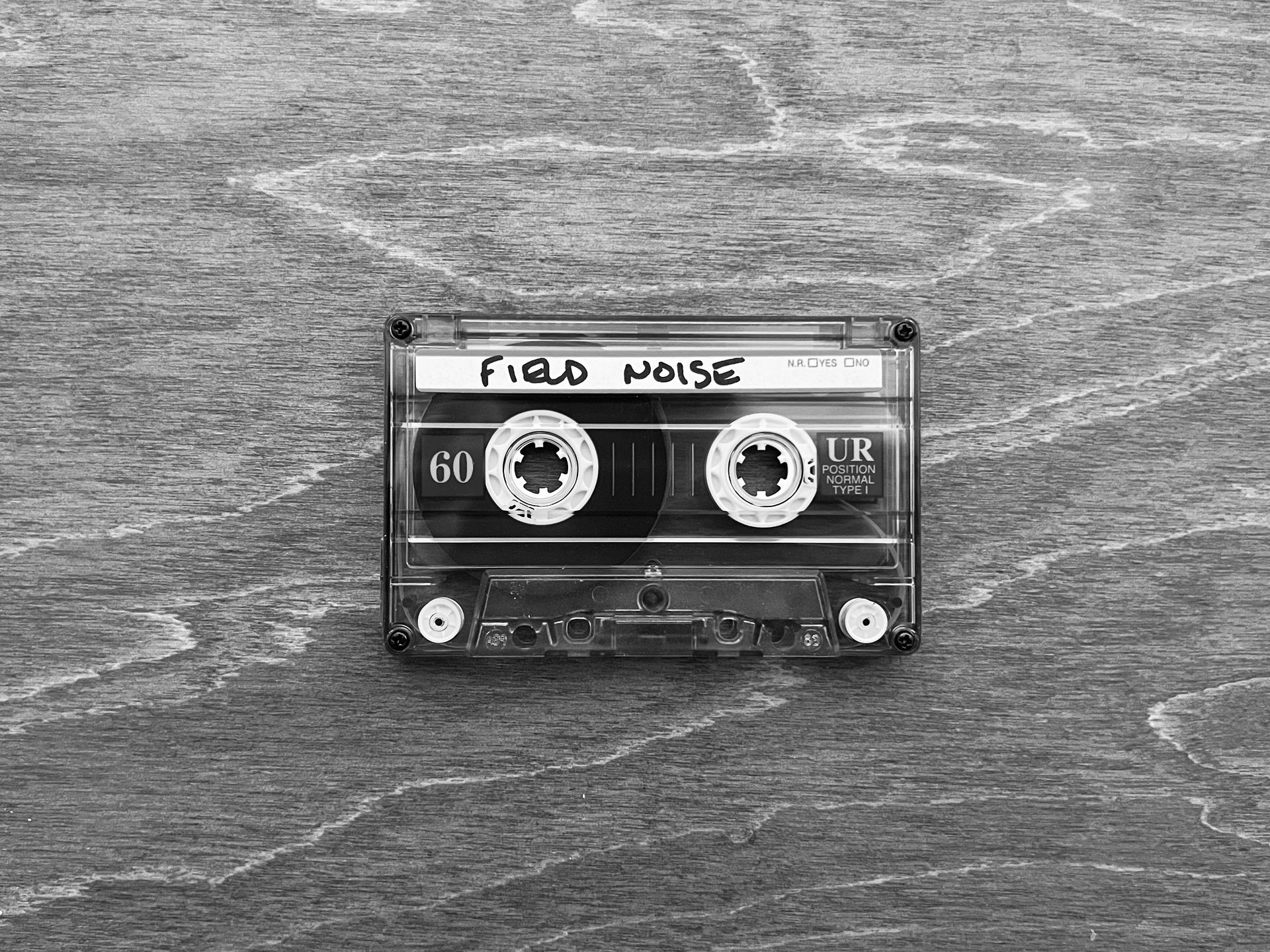 a black and white photo of a cassette tape labeled "field noise" sitting on a wooden desk.