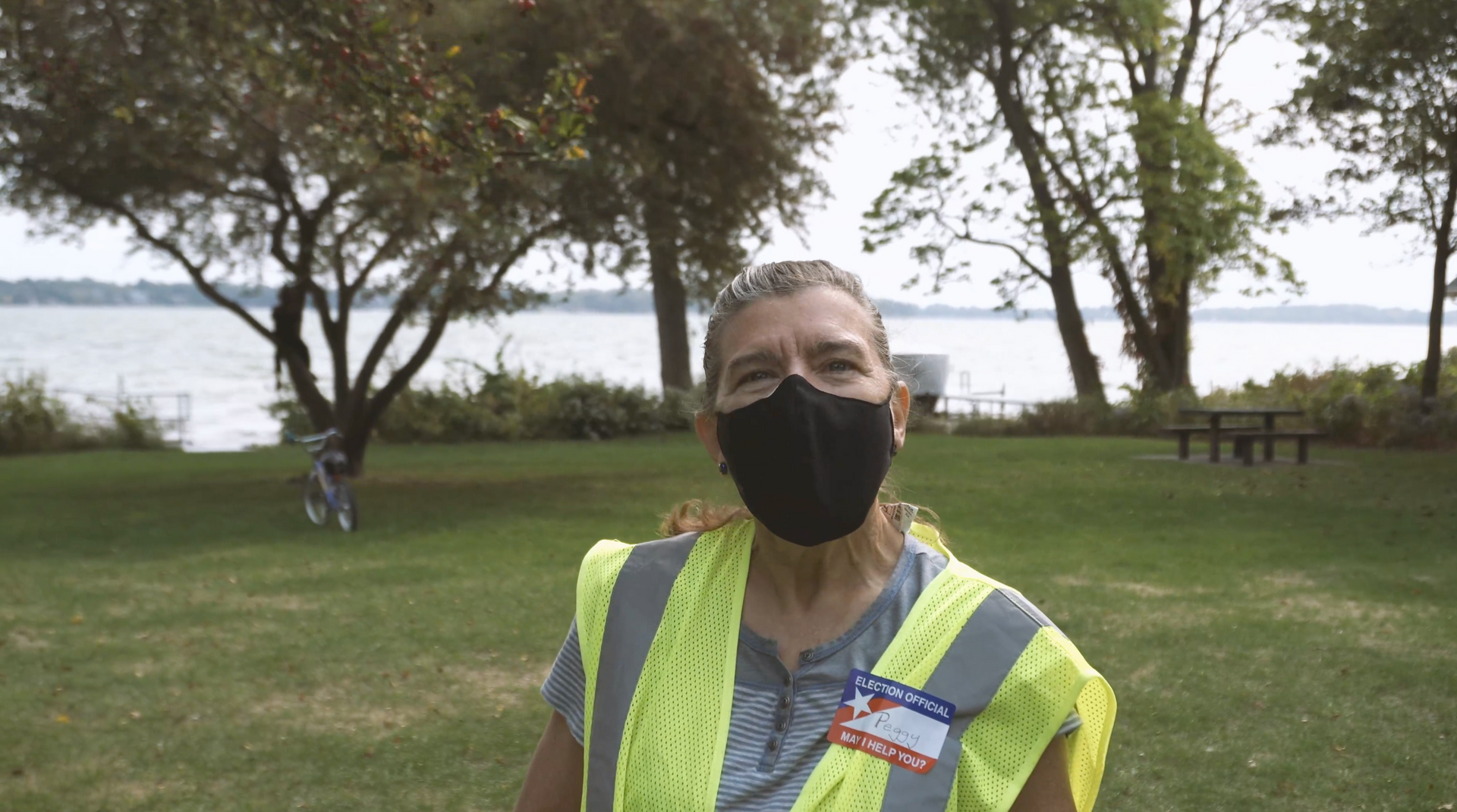 A woman in a yellow safety vest and black mask stands in a park and looks directly at the camera