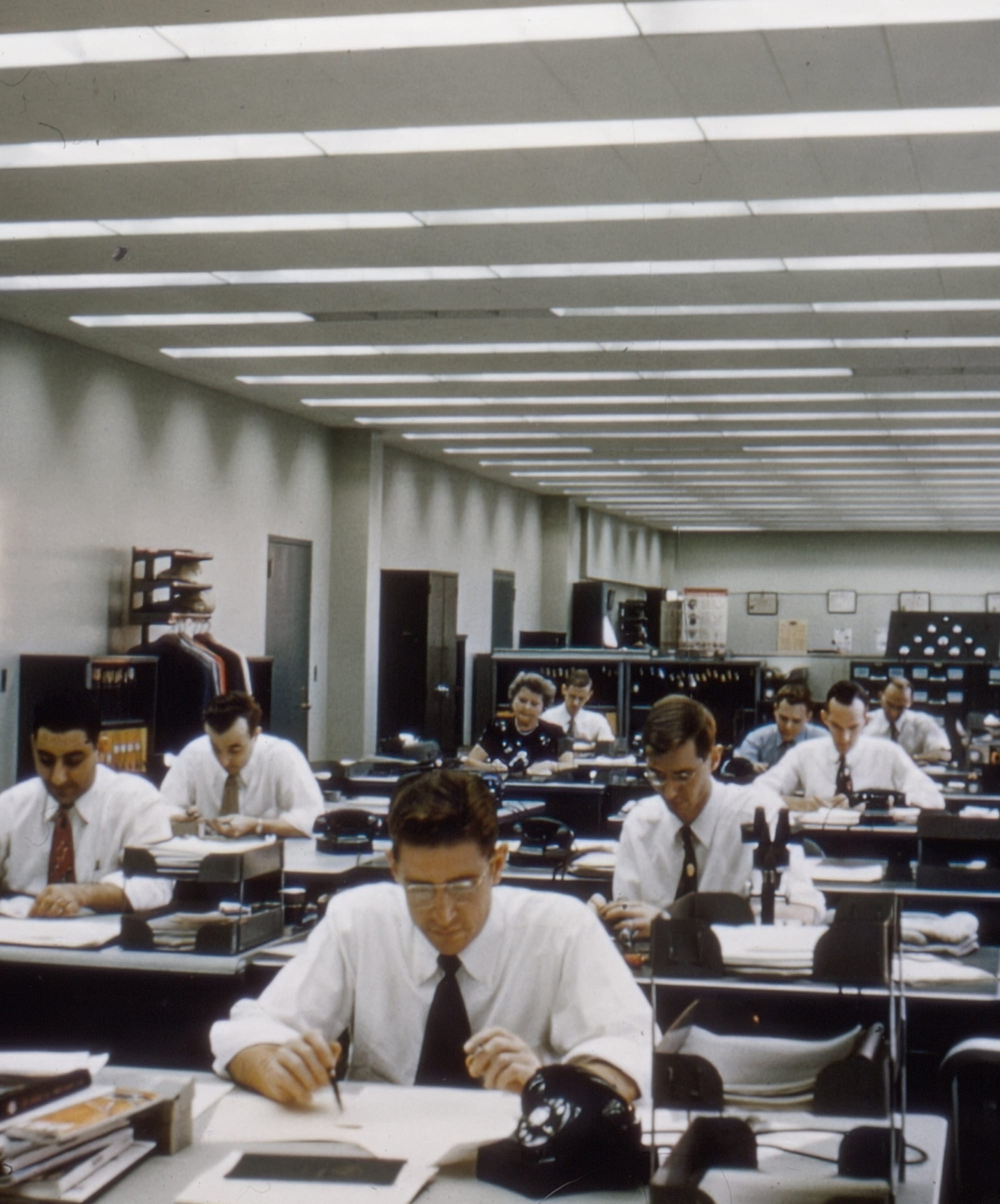 A group of people working in an open office in the late 1950s or early 1960s, all of them with their heads down looking at their desks.
